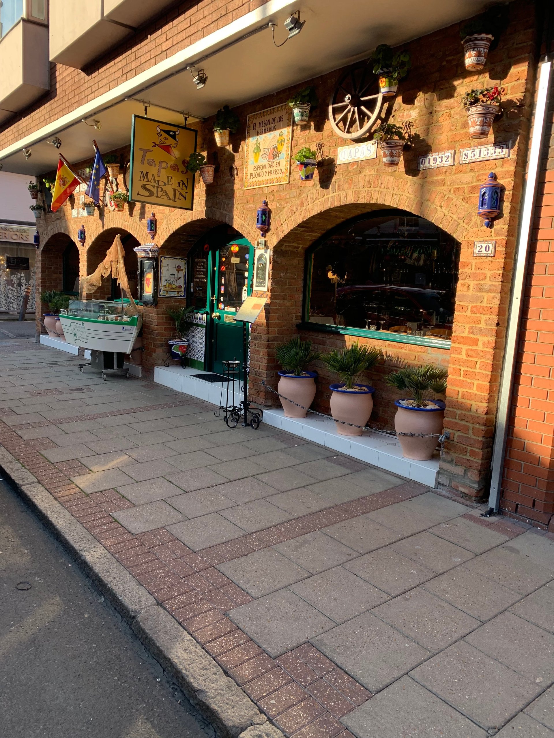 Brick storefront with arched windows, decorated with flags, hanging baskets, and potted plants on a tiled street